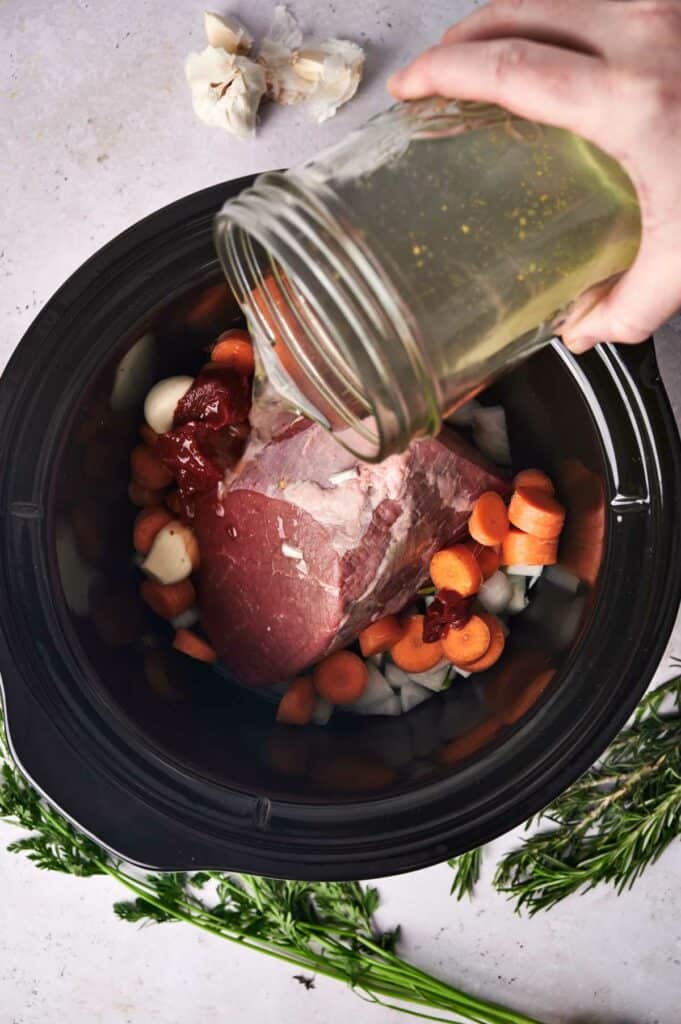 Preparing a slow cooker pot roast: pouring seasoning over beef and vegetables.