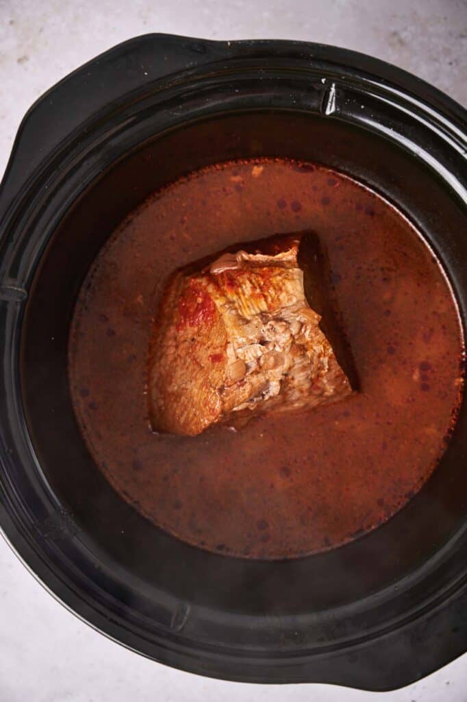 A piece of pot roast cooking in a slow cooker with juices.