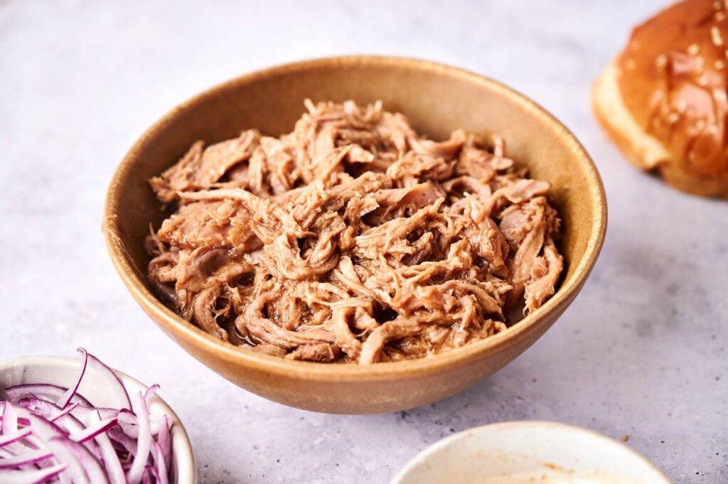 A brown bowl filled with Slow Cooker Pulled Pork sits on a light surface, with sliced red onions and part of a bun visible nearby.