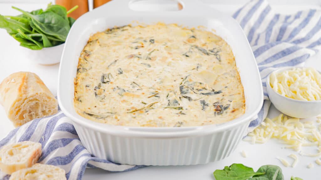 A spinach artichoke dip in a white baking dish, accompanied by bread rolls, shredded cheese, and fresh spinach leaves on the side.
