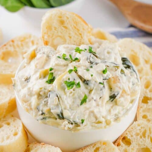 A bowl of creamy spinach artichoke dip served with slices of baguette and fresh spinach leaves in the background.