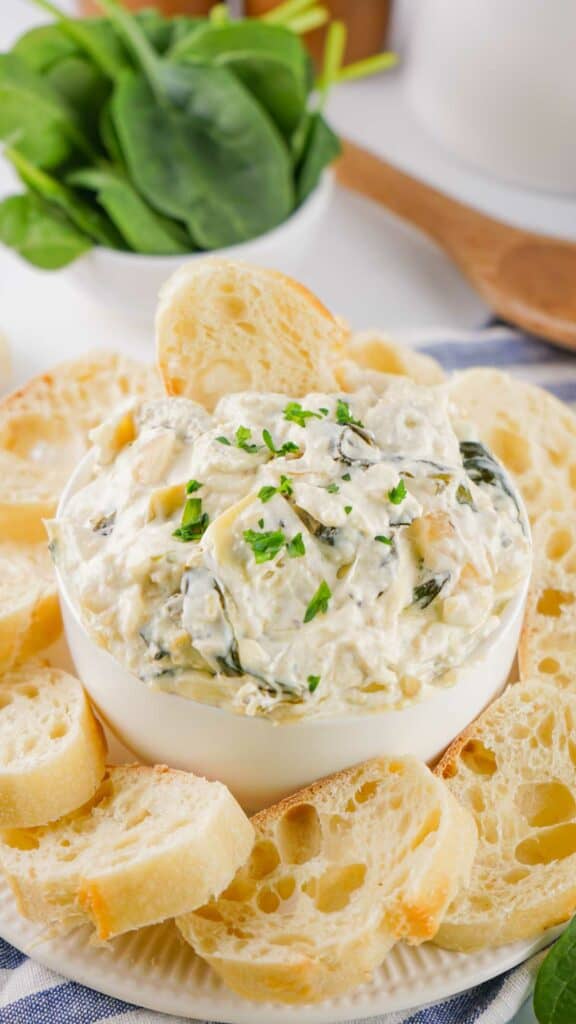 A bowl of creamy spinach artichoke dip served with slices of baguette and fresh spinach leaves in the background.