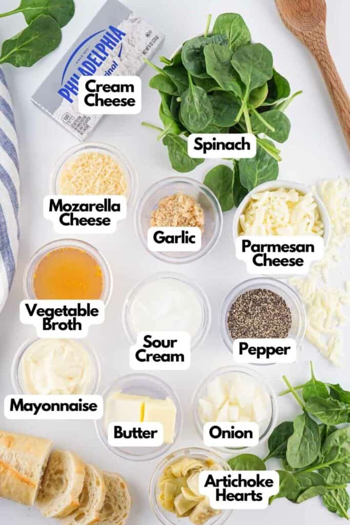 Ingredients for Spinach Artichoke Dip labeled on a white surface, including cream cheese, spinach, various cheeses, garlic, vegetable broth, sour cream, mayo, butter, and artichoke hearts