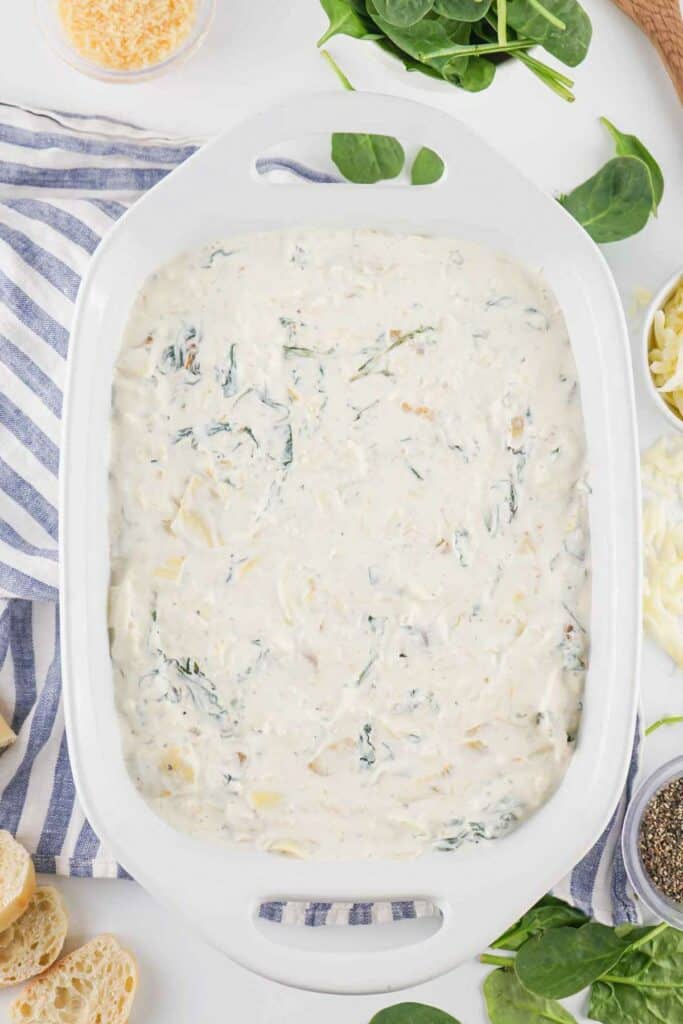 Spinach Artichoke Dip in a white ceramic dish on a striped cloth, surrounded by ingredients like cheese and bread.