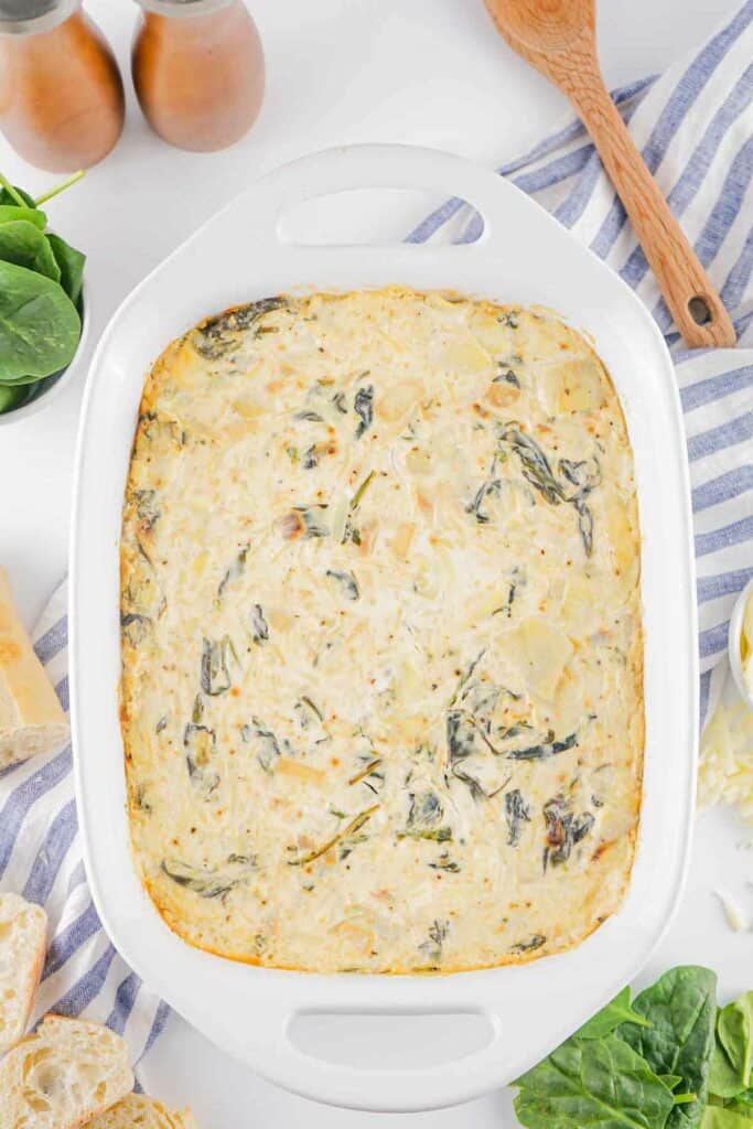 A creamy spinach artichoke dip with cheese in a white oval baking dish, with scattered fresh spinach leaves and slices of bread on a striped tablecloth.