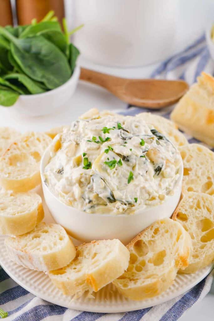 A creamy spinach artichoke dip with cheese in a white oval baking dish, with scattered fresh spinach leaves and slices of bread on a striped tablecloth.