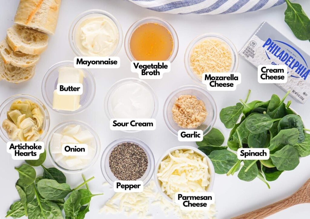 Overhead view of labeled ingredients for a Spinach Artichoke Dip, including mayonnaise, butter, sour cream, cream cheese, artichoke hearts, onion, pepper, vegetable broth, mozzarella, garlic, parmesan, and spinach.