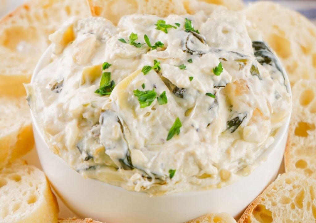 Creamy Spinach Artichoke Dip in a white bowl, garnished with chopped parsley and surrounded by slices of baguette. This classic appetizer is perfect for sharing at any gathering.
