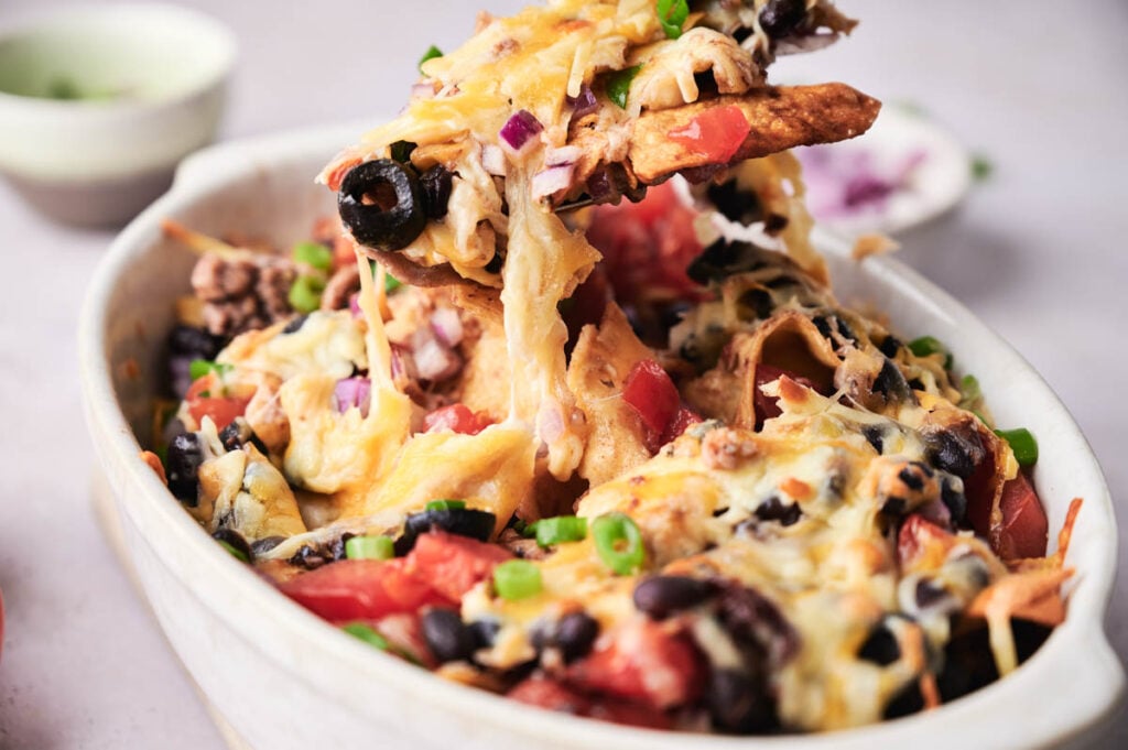 A dish of loaded nachos topped with melted cheese, black beans, tomatoes, olives, green onions, and red onions— the flavors of a classic taco casserole—served with a spoon lifting a portion.