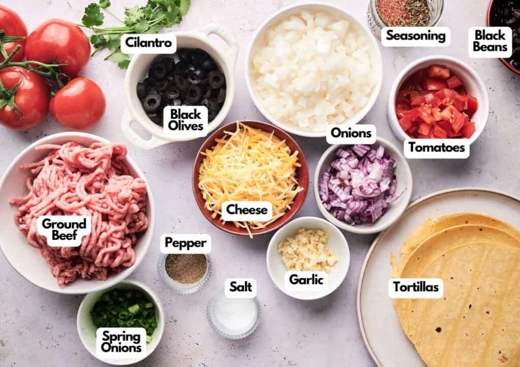A variety of taco ingredients are arranged on a counter, including ground beef, cheese, chopped vegetables, black beans, spices, and tortillas—perfect for making taco casserole—each labeled with text.
