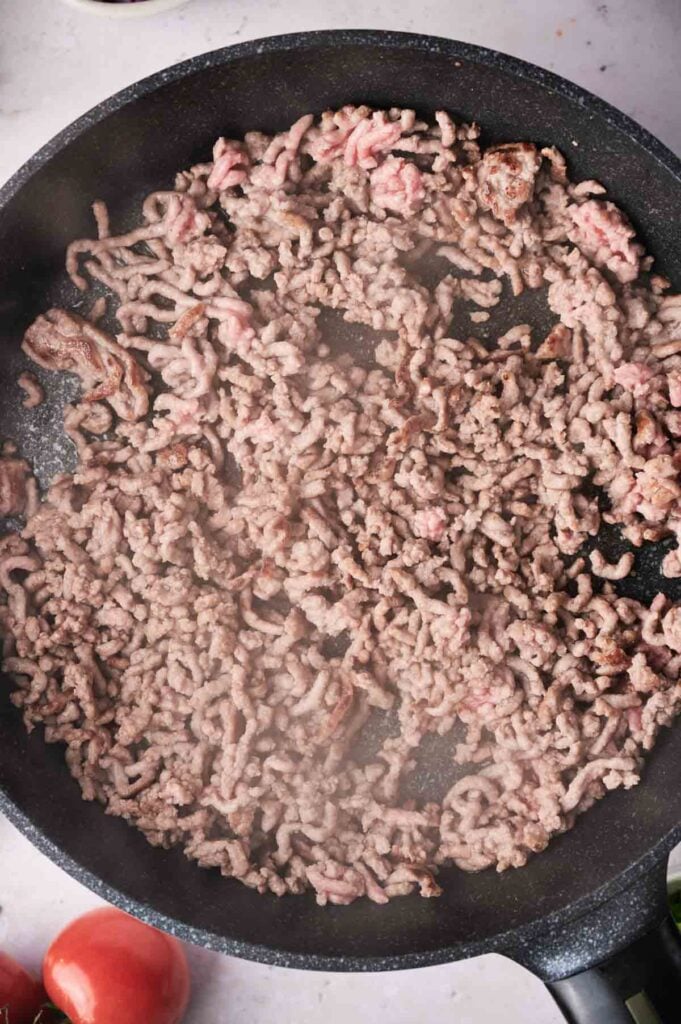 Ground beef cooking in a black skillet, with some pieces browned and others still pink—perfectly prepped for a flavorful taco casserole. Steam rises from the meat, hinting at the delicious meal to come.