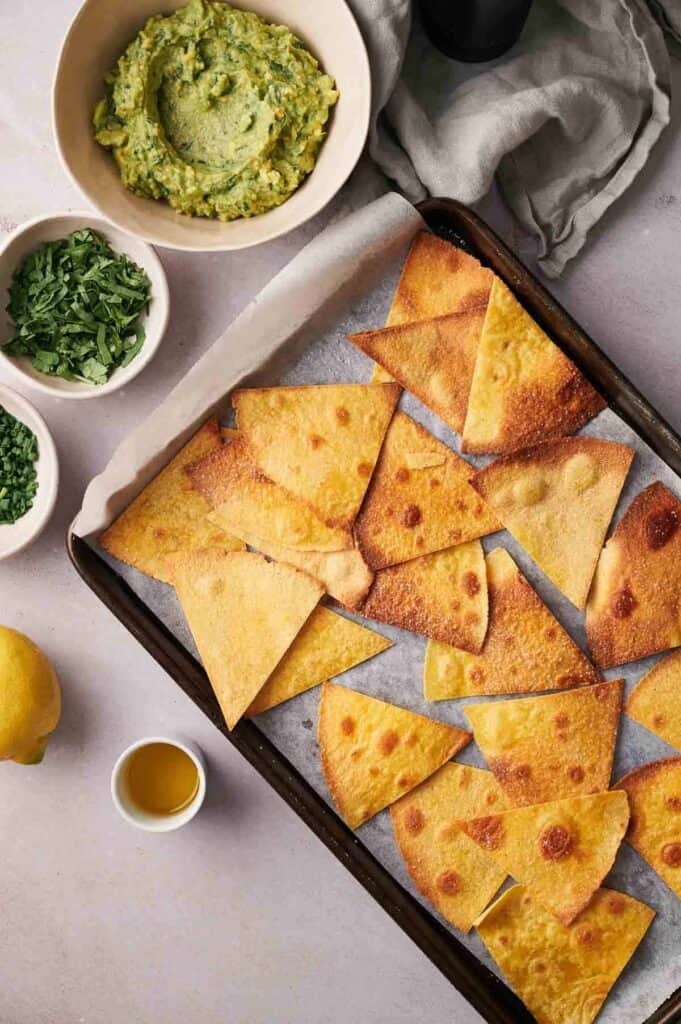 A tray of golden-brown pita chips paired with a bowl of green avocado dip, garnished with chopped herbs, lemon halves, and a small bowl of olive oil arranged on a light gray surface.
