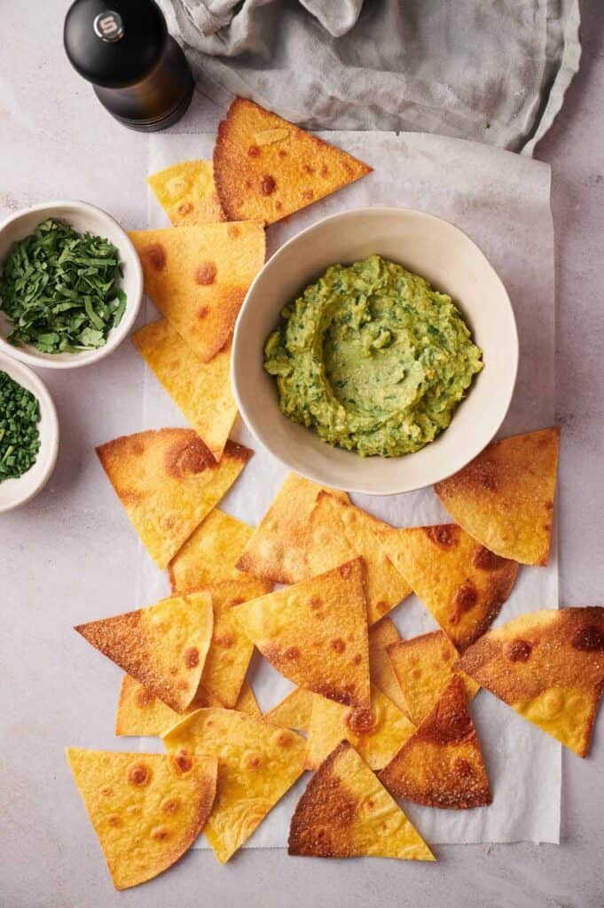 A bowl of guacamole surrounded by golden tortilla chips, with chopped herbs on the side and a beverage in the background on a light surface.