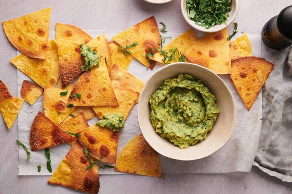 A bowl of guacamole surrounded by golden-brown tortilla chips scattered with green herbs on a light gray surface.