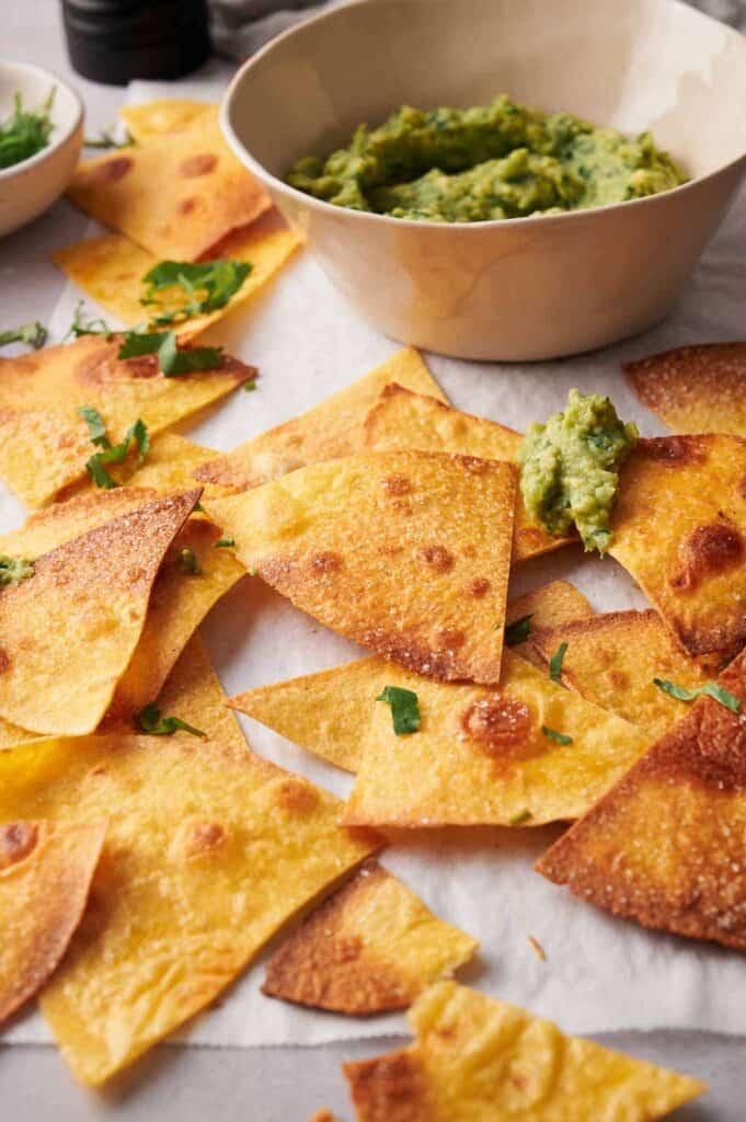 Homemade golden tortilla chips scattered around a bowl of guacamole, garnished with fresh cilantro on a light surface.