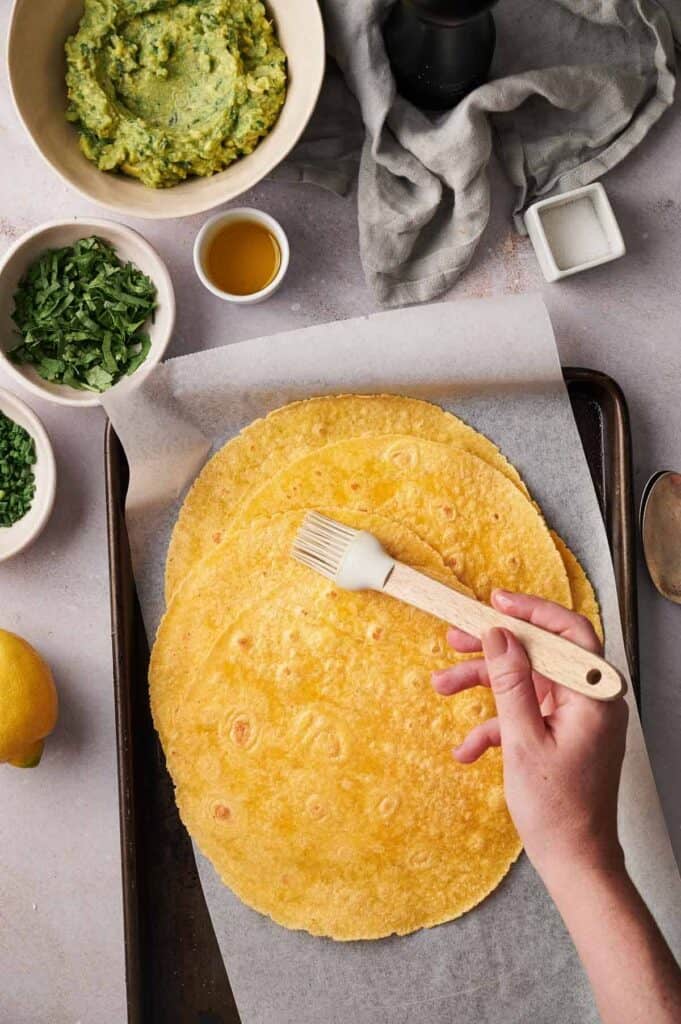 A person brushes oil on a large, round, golden tortilla on a baking sheet, with ingredients like guacamole and cilantro nearby.