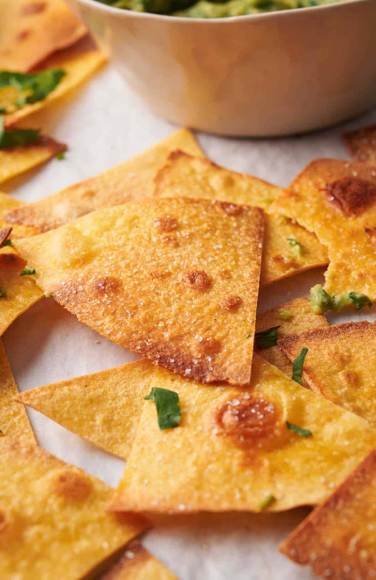 Golden-brown tortilla chips scattered on a surface, with chopped herbs sprinkled on top and a bowl of guacamole in the background.
