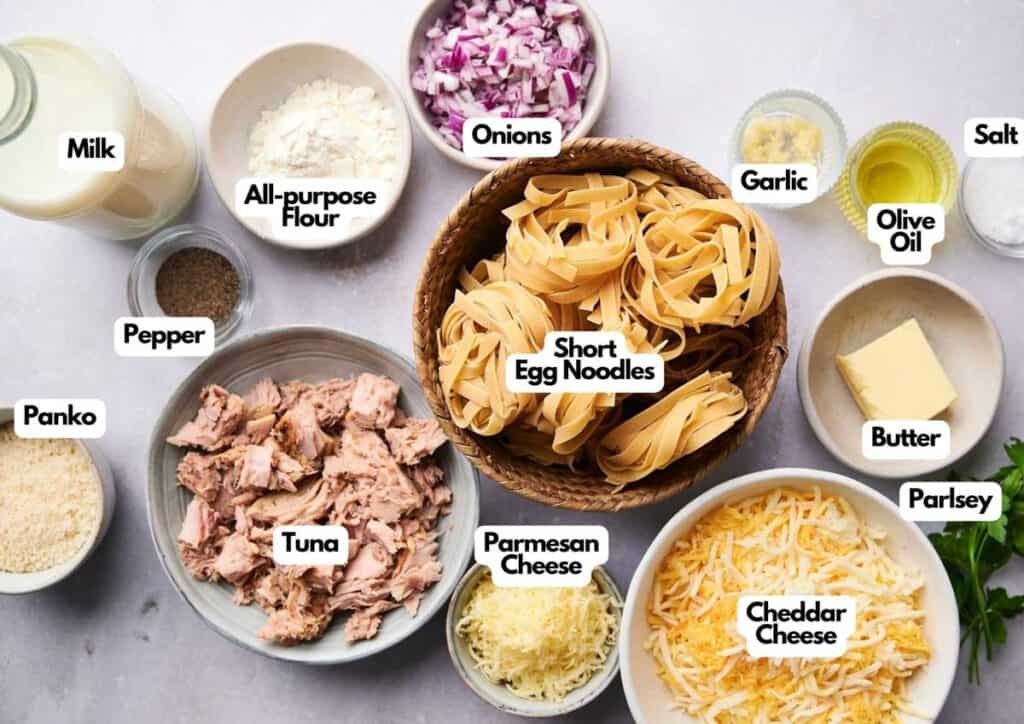 Ingredients for a tuna noodle casserole are arranged on a table, similar to how you&rsquo;d prep for a taco casserole, featuring milk, flour, onions, garlic, salt, olive oil, butter, egg noodles, panko, tuna, cheeses, pepper, and parsley.