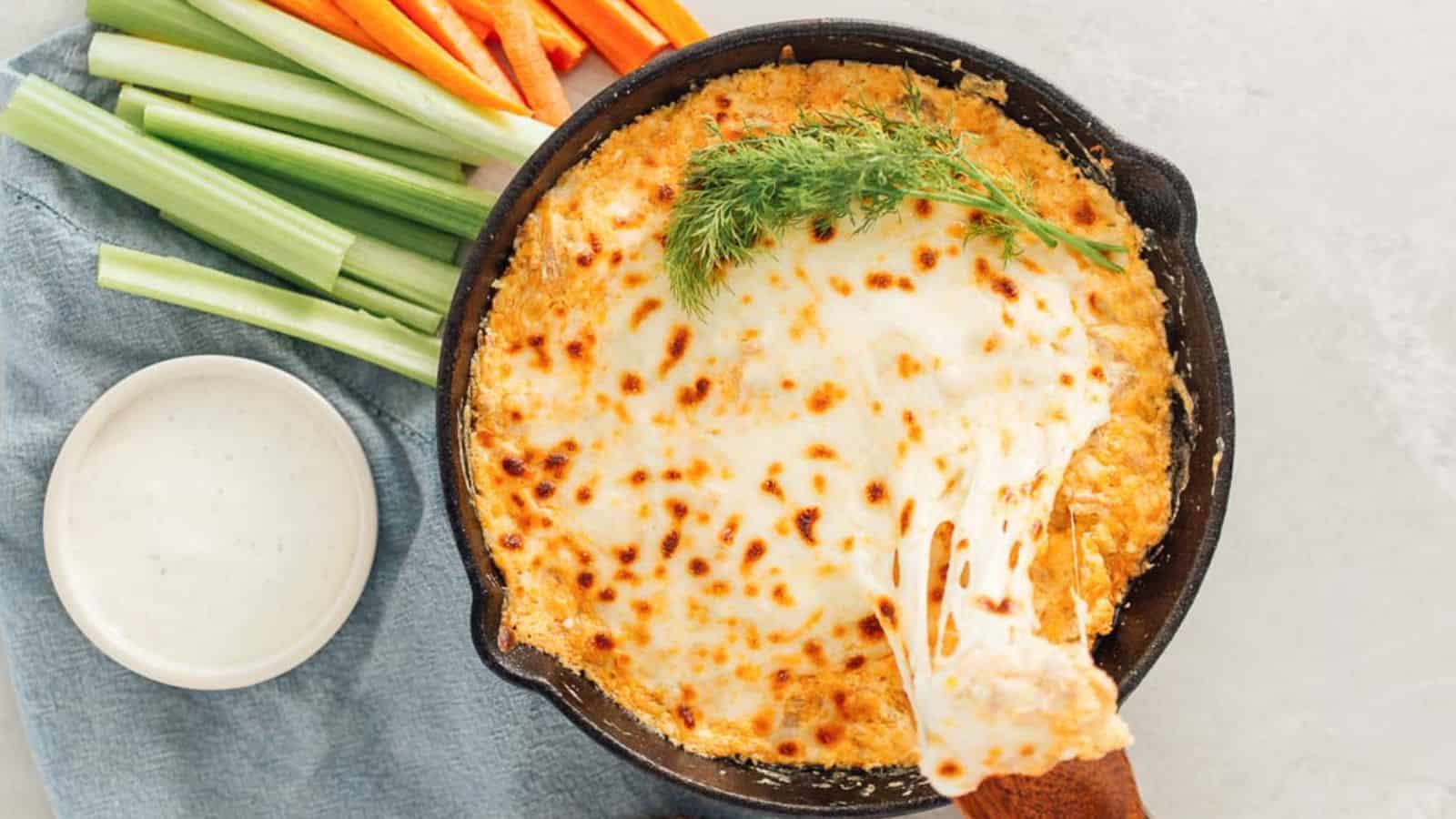 A skillet of bubbly, baked buffalo chicken dip with a piece being scooped out, served with celery and carrot sticks and a small bowl of ranch dressing.