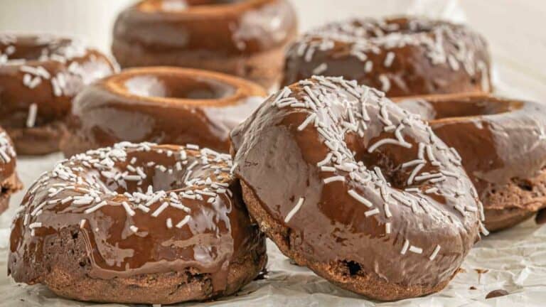 Chocolate glazed doughnuts topped with white sprinkles on parchment paper.