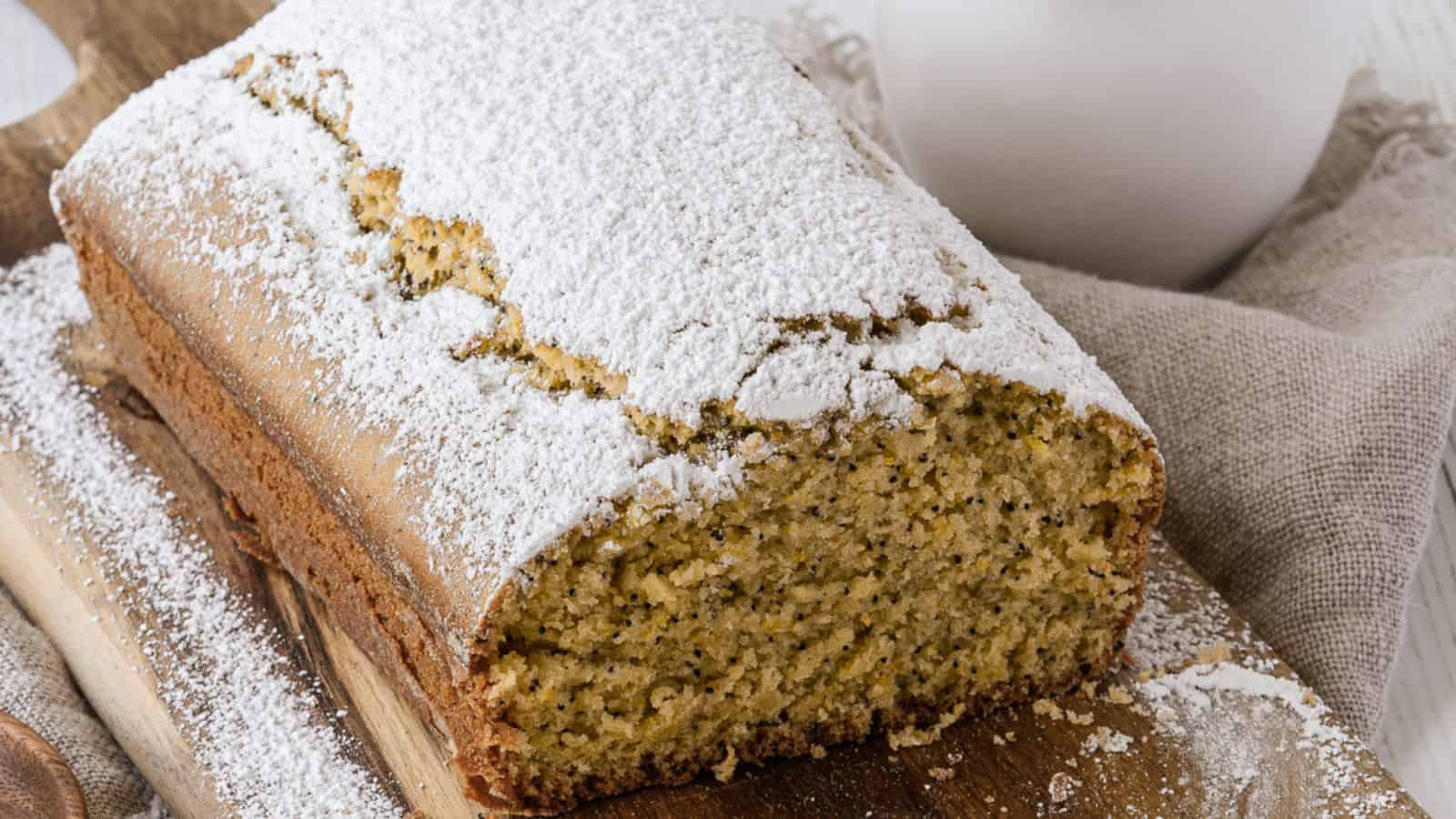 A loaf of poppy seed cake dusted with powdered sugar sits on a wooden board next to a beige cloth napkin.