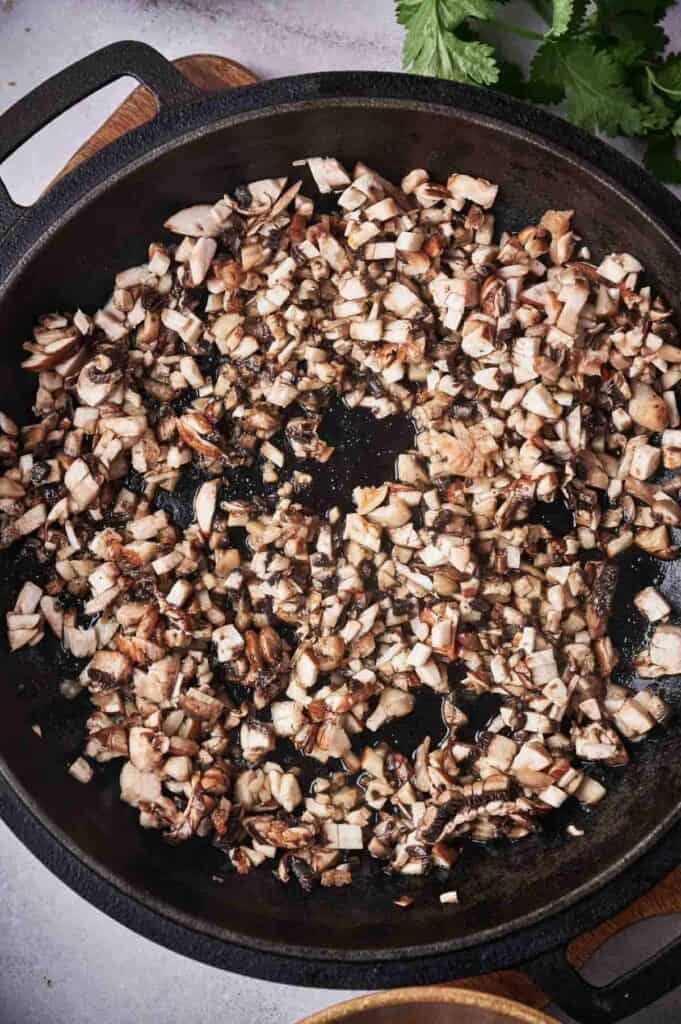 Diced mushrooms saut&eacute;ing in oil in a cast iron skillet, scattered with bits of chopped onions.