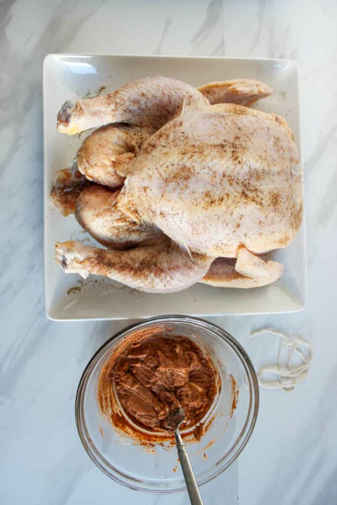 A whole raw chicken seasoned with spices next to a bowl of marinade on a kitchen countertop, ready to be made into baked bbq chicken.