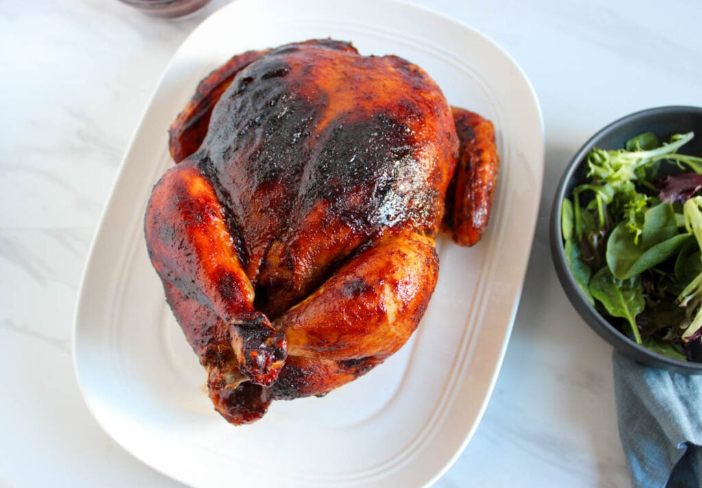 A whole baked BBQ chicken with dark, crispy skin is served on a white platter beside a bowl of mixed green salad.