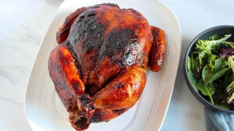 Whole baked BBQ chicken with a dark, glazed skin on a white serving platter sits next to a bowl of mixed green salad.