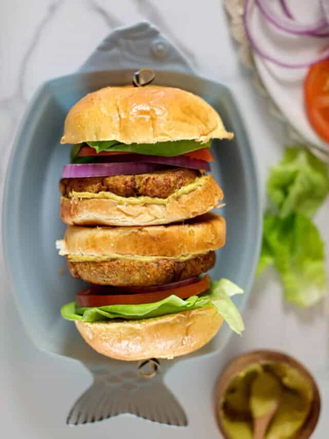 Two neatly stacked veggie burgers with lettuce, tomato, and red onion in buns placed on a fish-shaped plate. Sliced vegetables and a small bowl of pickles sit nearby.
