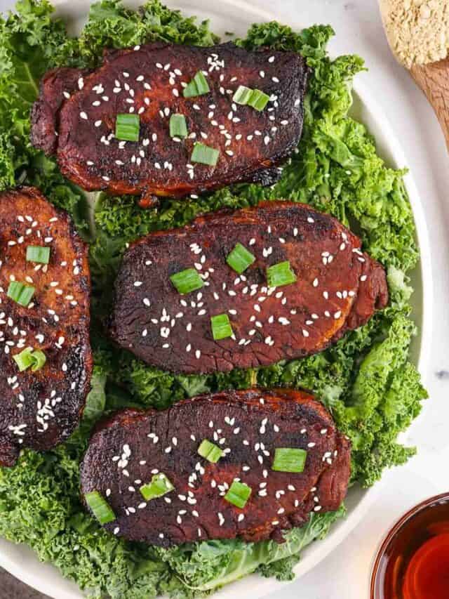 Four pieces of grilled meat, garnished with sesame seeds and chopped green onions, on a bed of leafy greens.