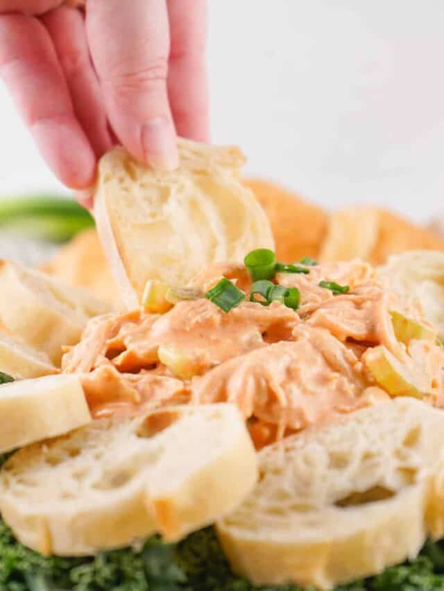 A hand dipping a slice of bread into a creamy dip garnished with chopped green onions, surrounded by additional bread slices.