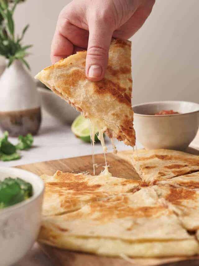 A hand lifting a slice of cheesy quesadilla, with stringy cheese visible, from a wooden board, with bowls of salsa and garnishes beside it.