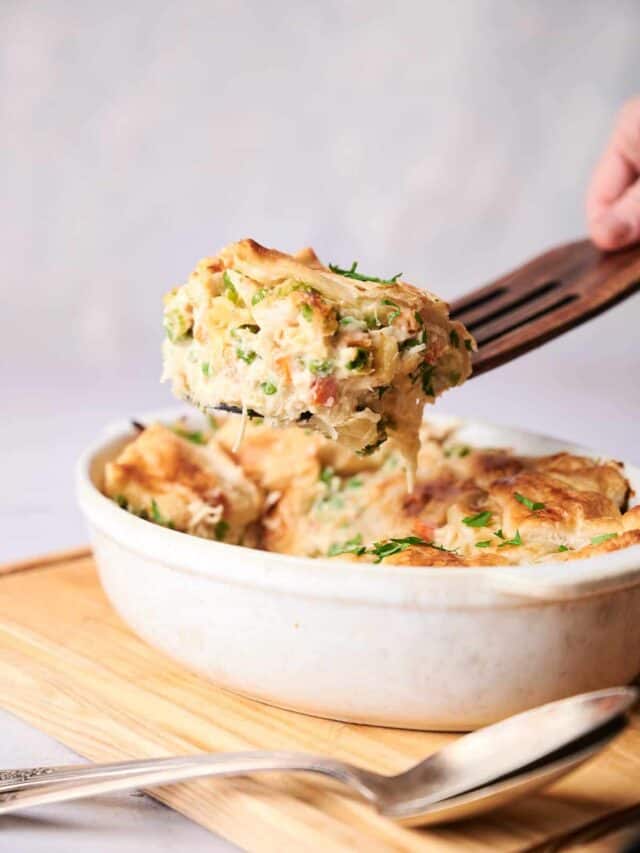 A slice of creamy chicken casserole is being served from an oval-shaped white dish resting on a wooden board, with a spoon placed beside it.