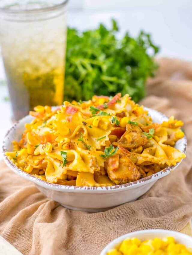 A bowl of creamy macaroni and cheese topped with chunks of grilled chicken and garnished with herbs, accompanied by a glass of iced tea and fresh greens in the background.