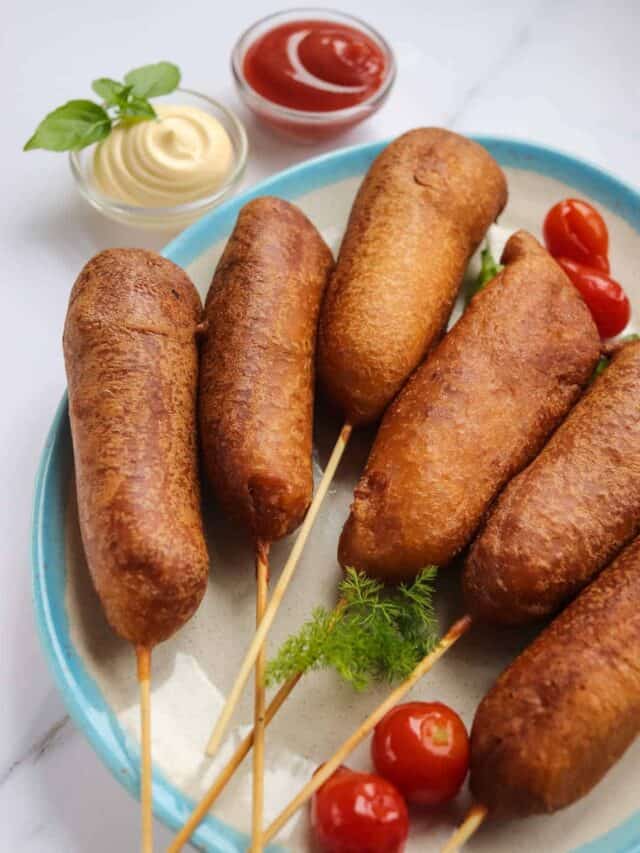 Five corn dogs on skewers served on a plate with ketchup, mayonnaise, cherry tomatoes, and fresh herbs.