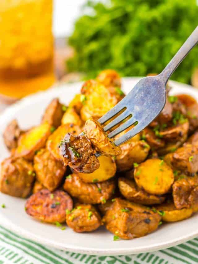 A fork holds up a piece of roasted potatoes garnished with herbs, above a plate filled with more roasted potatoes. In the background, there is a blurred image of greens and a beverage.