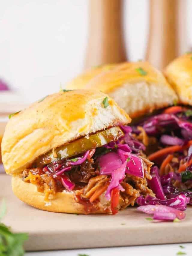 A pulled pork slider with pickles and a vibrant purple cabbage slaw on a wooden cutting board.