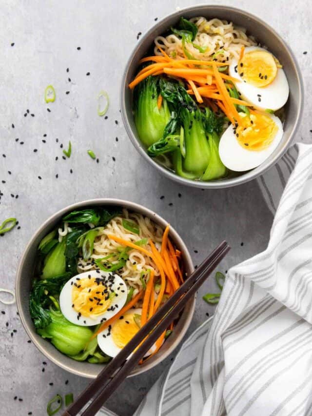 Two bowls of Asian noodle soup with boiled eggs, bok choy, carrots, and sesame seeds, garnished with green onions, served with chopsticks and a striped napkin.