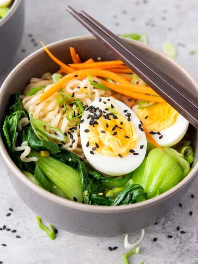 A bowl of ramen with vegetables, sliced egg, and sesame seeds, with chopsticks resting on the rim.