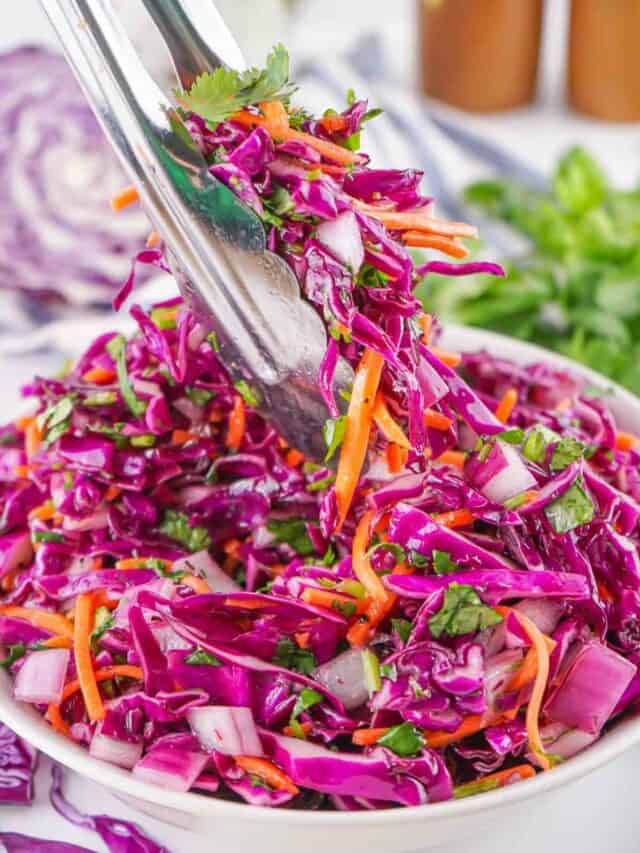A bowl of vibrant purple cabbage slaw with carrots, onions, and cilantro being tossed with tongs.