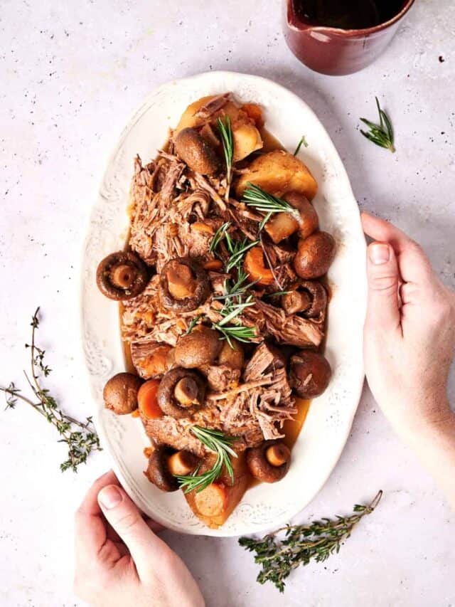 A platter of pulled meat and mushrooms garnished with rosemary, with hands adjusting the presentation.