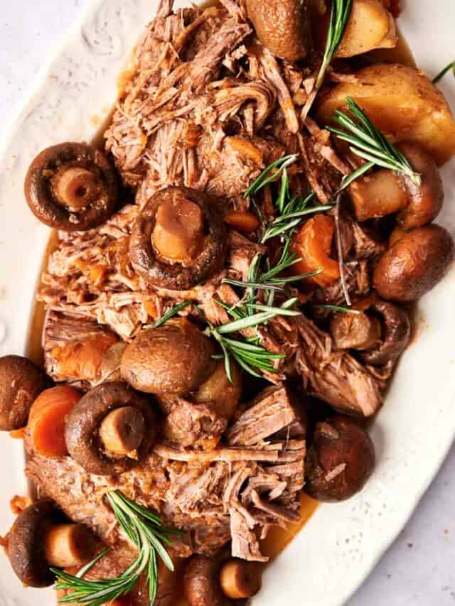Pot roast with carrots, mushrooms, and potatoes garnished with rosemary sprigs on a white plate.