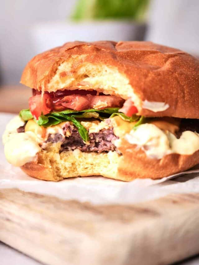 A close-up of a partially eaten cheeseburger with lettuce, tomato, and toppings on a wooden surface.