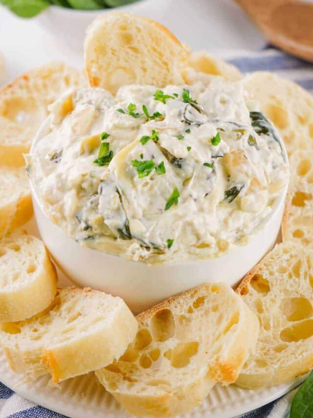 A bowl of creamy spinach artichoke dip garnished with parsley and surrounded by sliced baguette pieces on a plate.