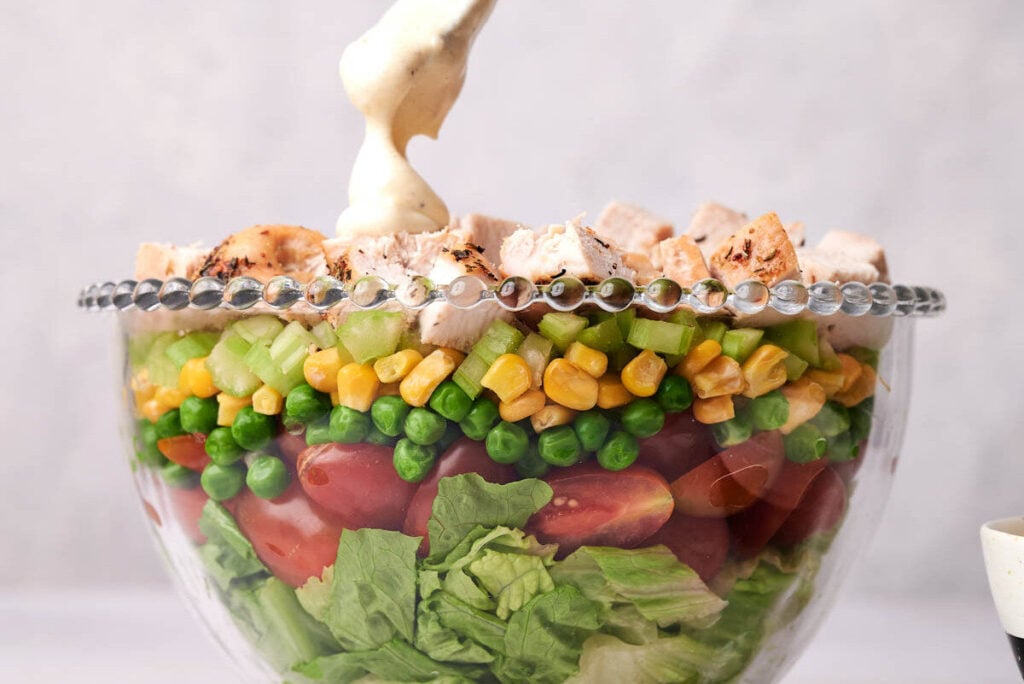 A glass bowl with layered salad ingredients—lettuce, tomatoes, peas, corn, celery, chicken—topped with a spoonful of creamy dressing creates a classic Seven Layer Salad.