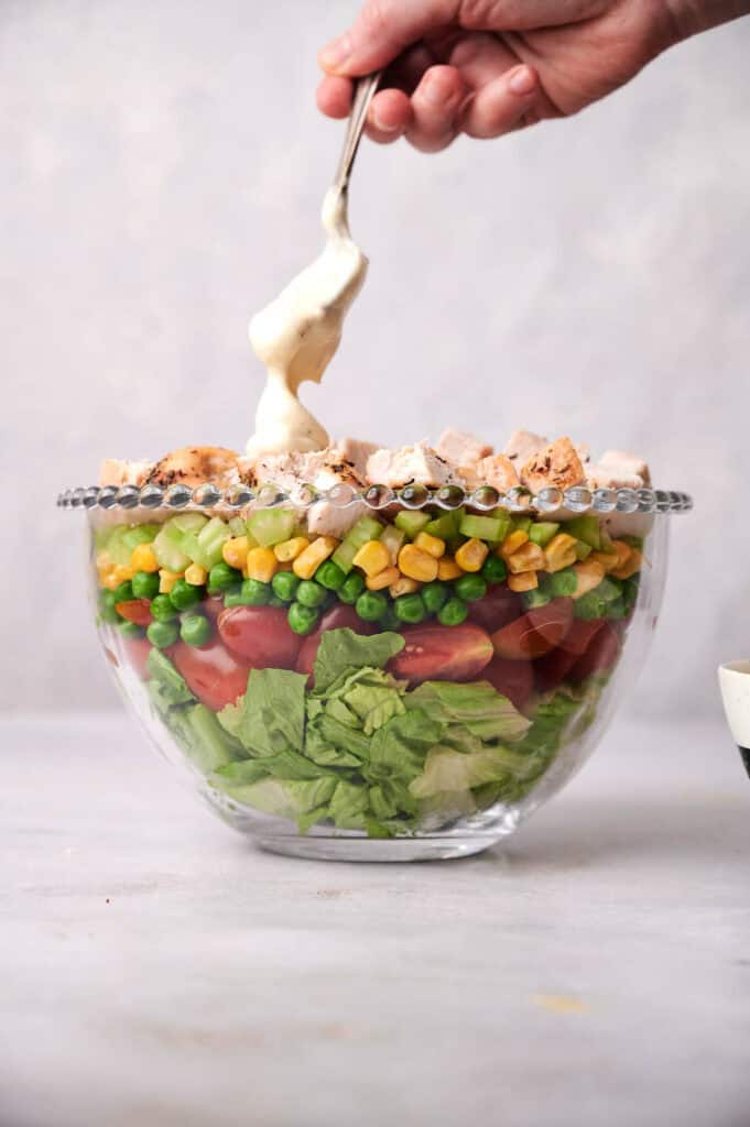 A hand pours dressing onto a beautifully presented Seven Layer Salad in a clear glass bowl, featuring layers of lettuce, peas, cherry tomatoes, corn, and chopped chicken.
