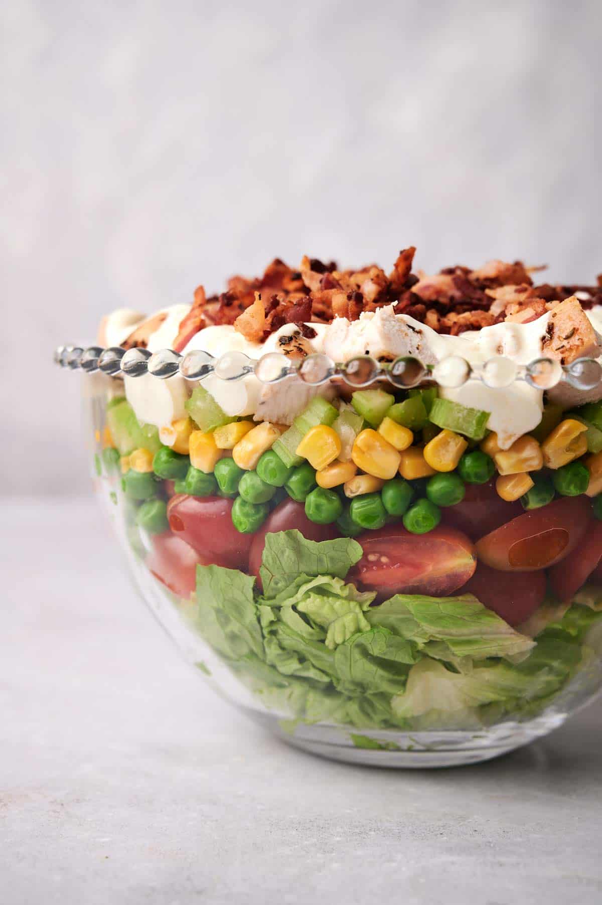 A glass bowl filled with a Seven Layer Salad including lettuce, tomatoes, peas, corn, cucumbers, cheese, and topped with bacon bits.