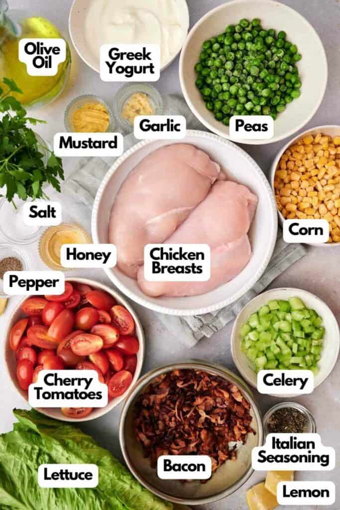 A top-down view of a Seven Layer Salad featuring labeled ingredients: chicken breasts, Greek yogurt, garlic, peas, corn, celery, Italian seasoning, lemon, honey, mustard, salt, pepper, cherry tomatoes; bacon and lettuce.