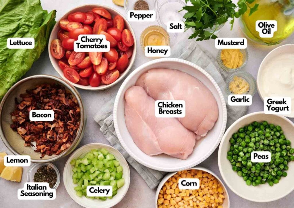 Ingredients for a classic Seven Layer Salad are arranged in bowls, featuring chicken breasts, bacon, cherry tomatoes, lettuce, celery, peas, corn, garlic, Greek yogurt, and assorted seasonings.